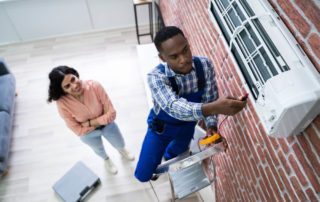 Young Woman Looking at Male Technician Repairing Air Conditioner Mounted on Brick Wall