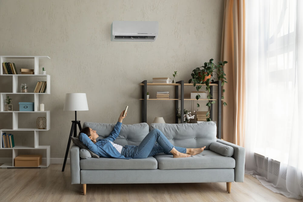 Glad Millennial Female Buyer of Air Conditioner Relax on Sofa Hold Controller Breath Cool Fresh Air Despite of Hot Summer Day Outside. 