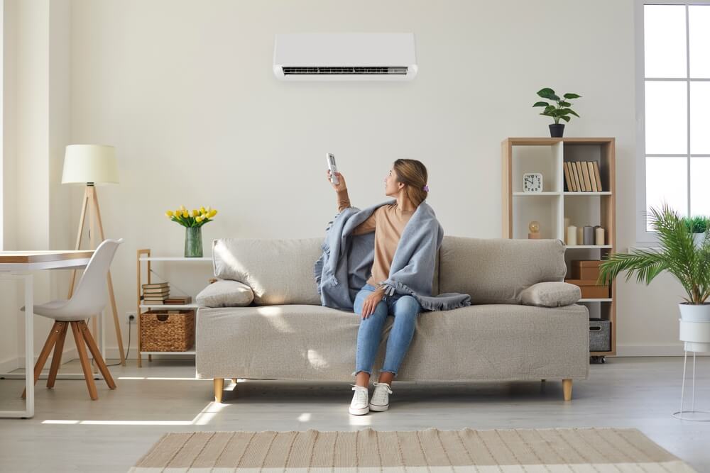 Woman Who’s Sitting on Sofa Under Warm Plaid in Living Room Switches off Her Air Conditioner on Wall.