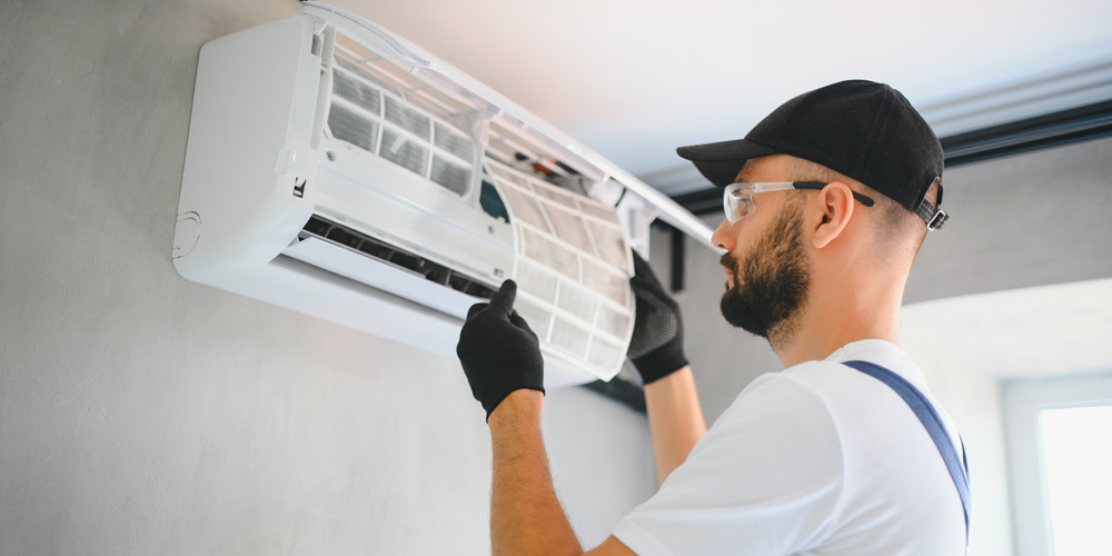 A technician wearing a black cap gloves and safety glasses services a wall-mounted air conditioner | Chills AC