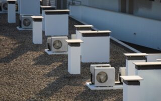 Rooftop with multiple white HVAC units installed on a gravel surface beside a pale building wall.