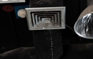 Square metal ceiling vent with concentric rectangular frames, water droplets below, beside a reflective lamp in a dark industrial ceiling area