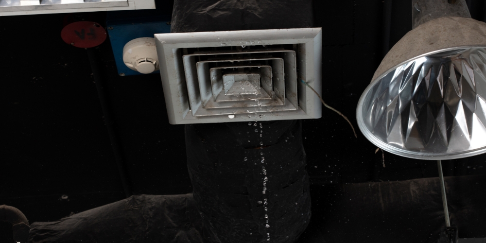 Square metal ceiling vent with concentric rectangular frames, water droplets below, beside a reflective lamp in a dark industrial ceiling area