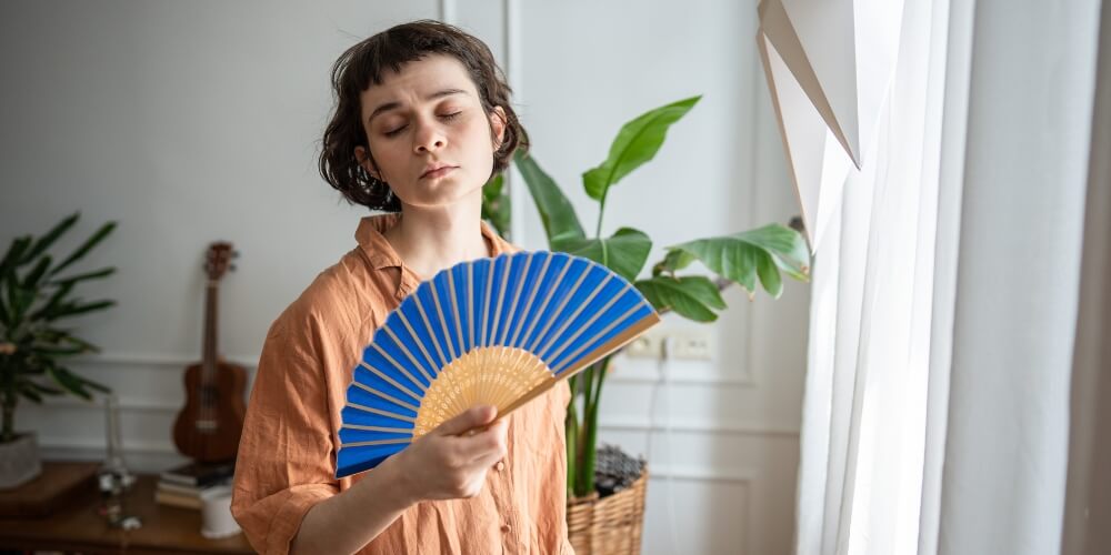 A woman with a fan poses by a window | Chills AC Woman with eyes closed gently fanning herself with a blue handheld fan indoors, near plants and a guitar in the background.