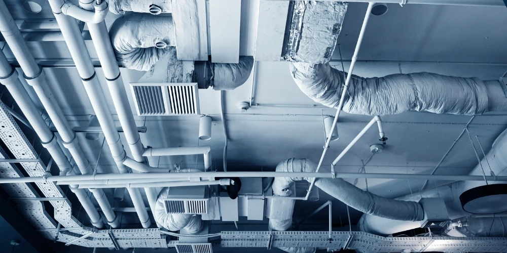 A room filled with pipes | Chills AC Industrial ceiling with a dense network of insulated pipes, ducts, and vents in a mechanical room (blue tint)