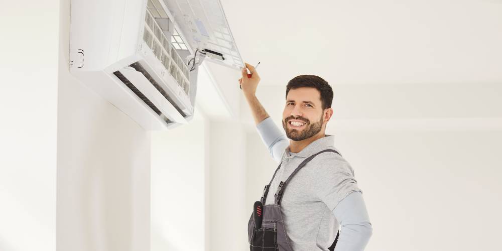 Salt Air Corrosion 2 | Chills AC HVAC technician wearing gray shirt and work overalls adjusting a wall-mounted split air conditioner, smiling at the camera.