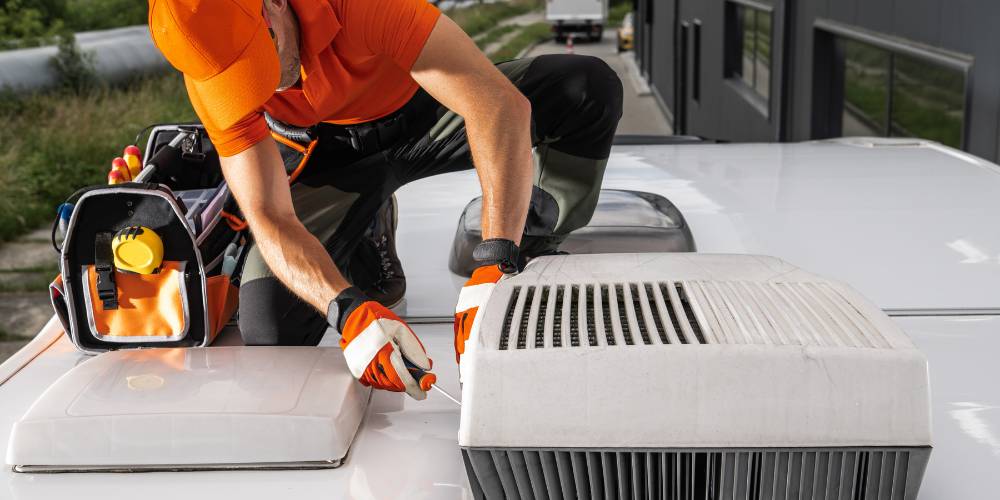air conditions unit repair | Chills AC HVAC technician in an orange uniform and gloves kneels on a roof, using a screwdriver beside a tool bag to service the outdoor air conditioning unit.
